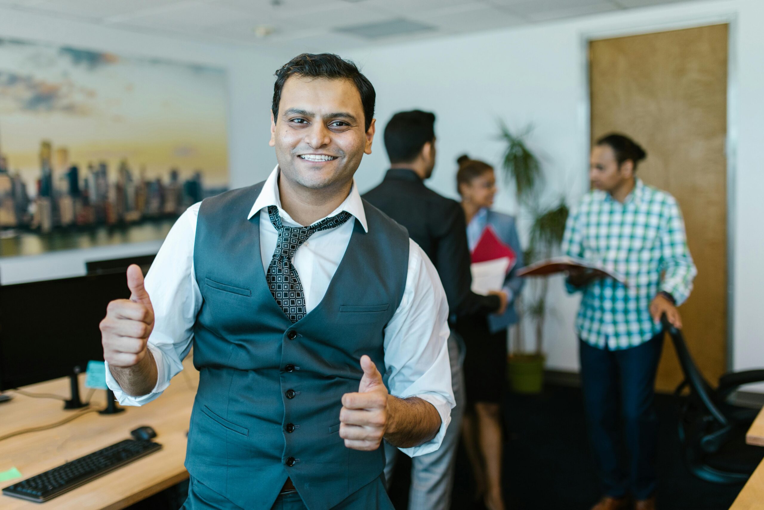 Smiling businessman showing thumbs up in a lively office environment with colleagues in the background.